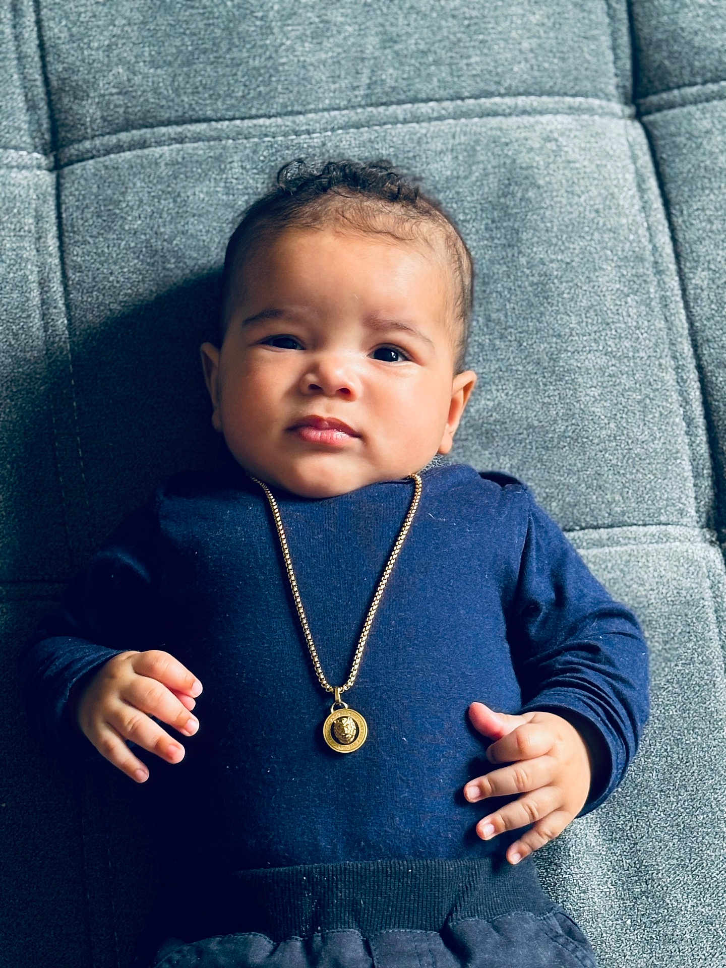 Yaya participe au concours pour gagner de l'argent avec cette photo : baby, infant, child, face, portrait, clothing, necklace, gold_jewelry, blue_shirt, hand, fingers, lying_down, texture, gray_background, cute, expression, young, skin, person, indoors