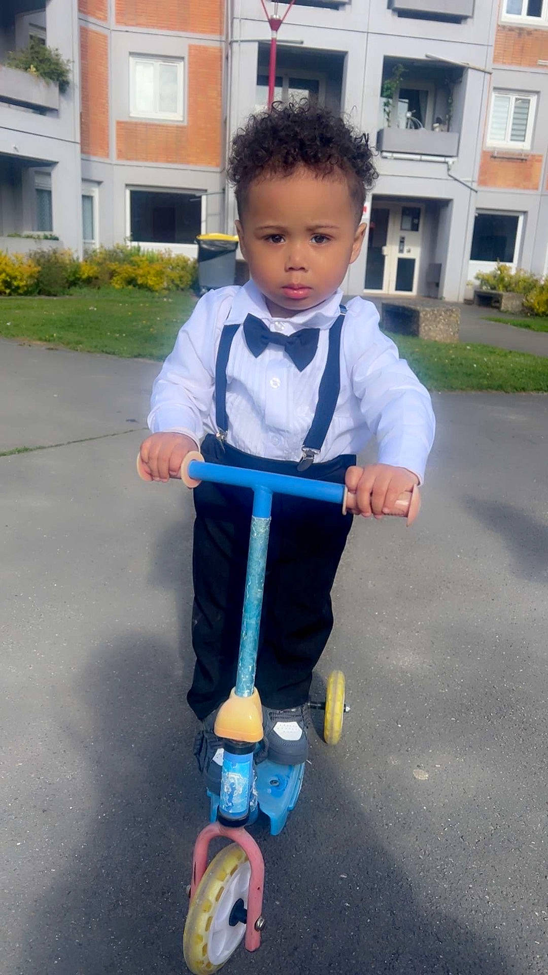 Tidiane participe au concours pour gagner de l'argent avec cette photo : bow_tie, boy, building, child, curly_hair, formal_clothing, grass, outdoor, pavement, person, play, residential_area, scooter, serious_expression, shoes, sunny_day, suspenders, toddler, vehicle, window