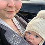 adult, baby, blue_eyes, knitted_hat, pom_pom, baby_carrier, smile, outdoor, car, close_up, face, person, winter_clothing, warm, curious, portrait, headwear, parent, child, cozy