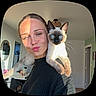 woman, cat, siamese_cat, pet, portrait, selfie, face, blue_eyes, duck_face, shoulder, black_sweater, indoor, home, room, furniture, door, window, sunlight, whiskers, playful
