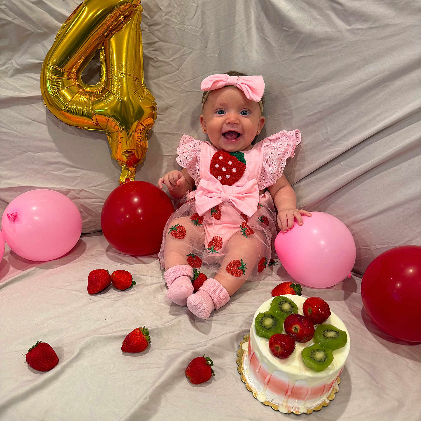 Maryory is registered to the contest to win money with this photo: baby, child, birthday, birthday_party, pink_bow, pink_outfit, strawberries, kiwi, cake, balloons, gold_number_four, infant, smiling, sitting, indoors, bed, blanket, fruit, dessert, portrait