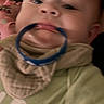 baby, child, face, mouth, ring, bib, green_shirt, pink_blanket, blur, closeup, infant, person, clothing, indoor, portrait, couch, head, young_child, baby_accessory, lying_down