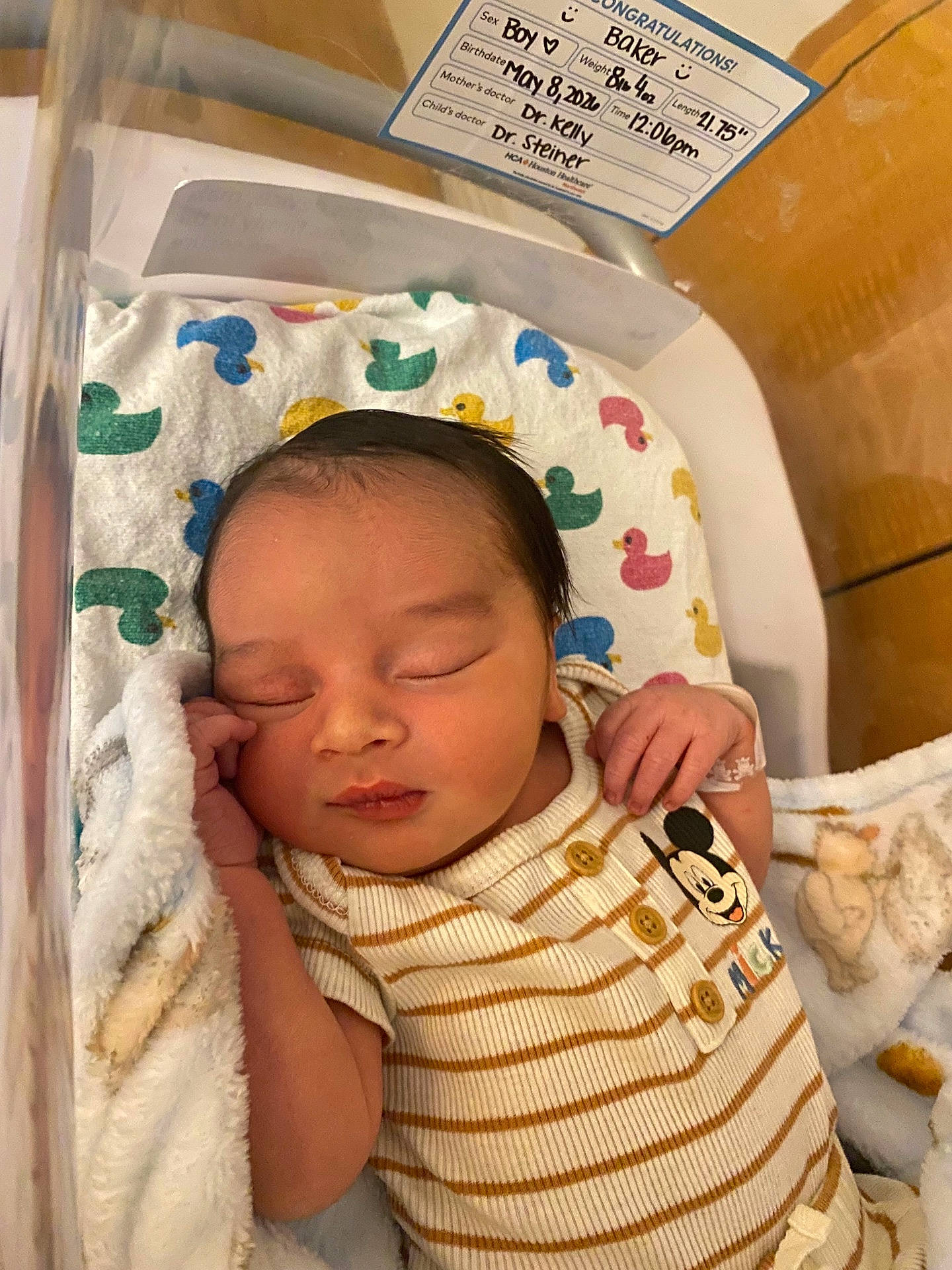 baby, bassinet, birth_info, blanket, child, closed_eyes, cozy, cute, duck_pattern, face, hand, hospital, hospital_card, infant, mickey_mouse, newborn, resting, sleeping, soft_toy, striped_onesie