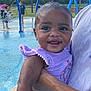 baby, child, smiling, water_park, splash_pad, outdoor, purple_clothing, wet_hair, person, holding, summer, play, fountain, water, happy, cute, toddler, daylight, people, park