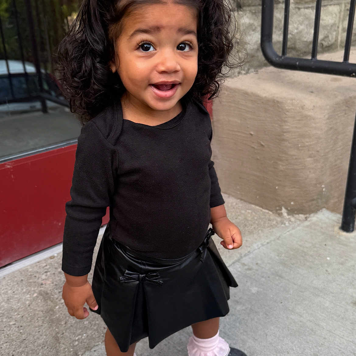 Azariah joined the competition — help win amazing prizes! black_crocs, black_skirt, black_top, child, curly_hair, cute, door, fashion, happy, outdoor, person, pink_bows, pink_socks, portrait, sidewalk, smiling, standing, stone_wall, toddler, young_child