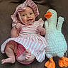Penelope is registered to the contest to win money with this photo: baby, child, infant, pink_clothing, striped_dress, bonnet, smiling, happy, face, feet, hands, stuffed_animal, duck, plush_toy, soft_texture, brown_background, lying_down, cute, adorable, indoor