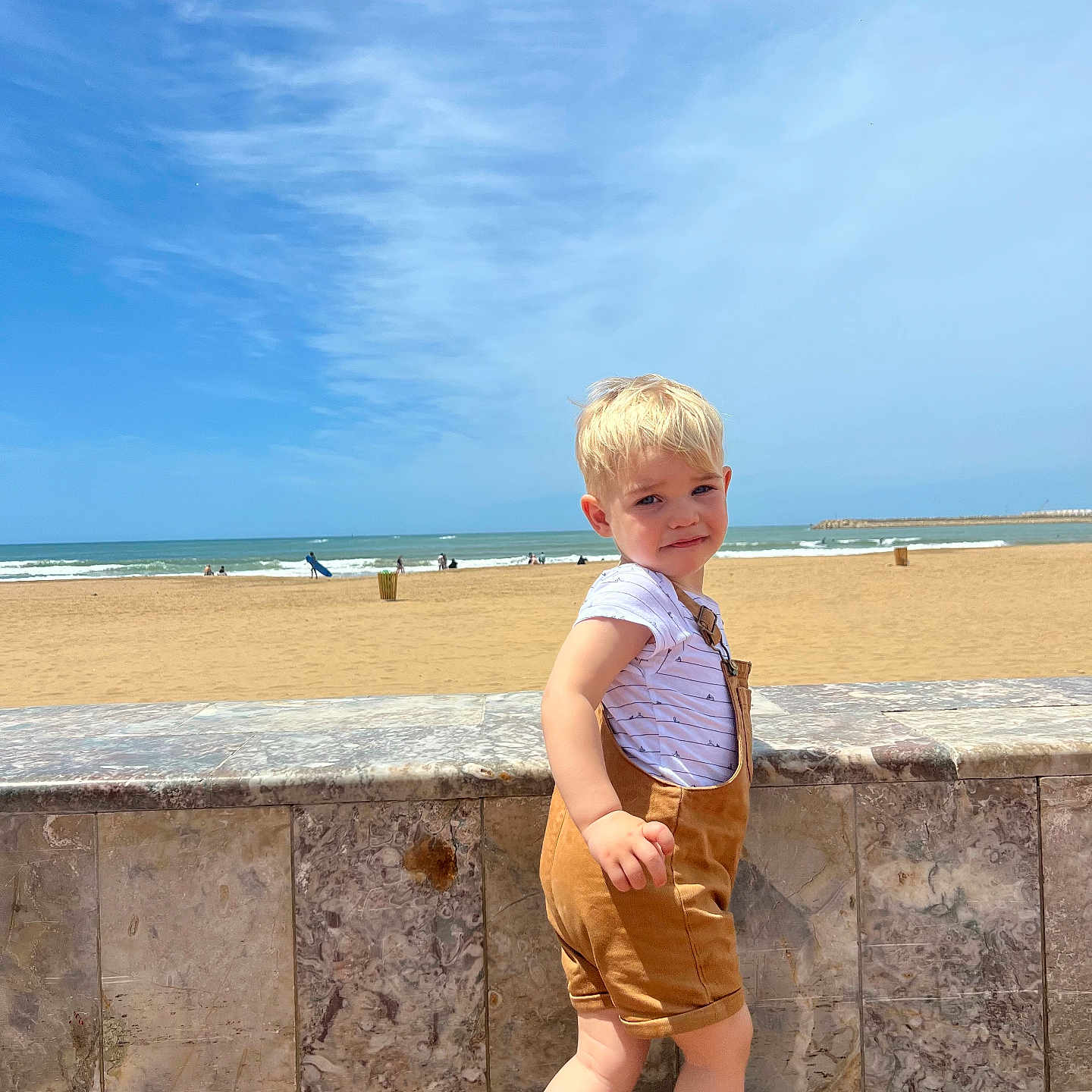 Milann participe au concours pour gagner de l'argent avec cette photo : beach, blond, casual, child, clouds, cute, footwear, marble_wall, ocean, outdoor, overalls, person, portrait, sand, sky, sneakers, standing, striped_shirt, sunny, toddler