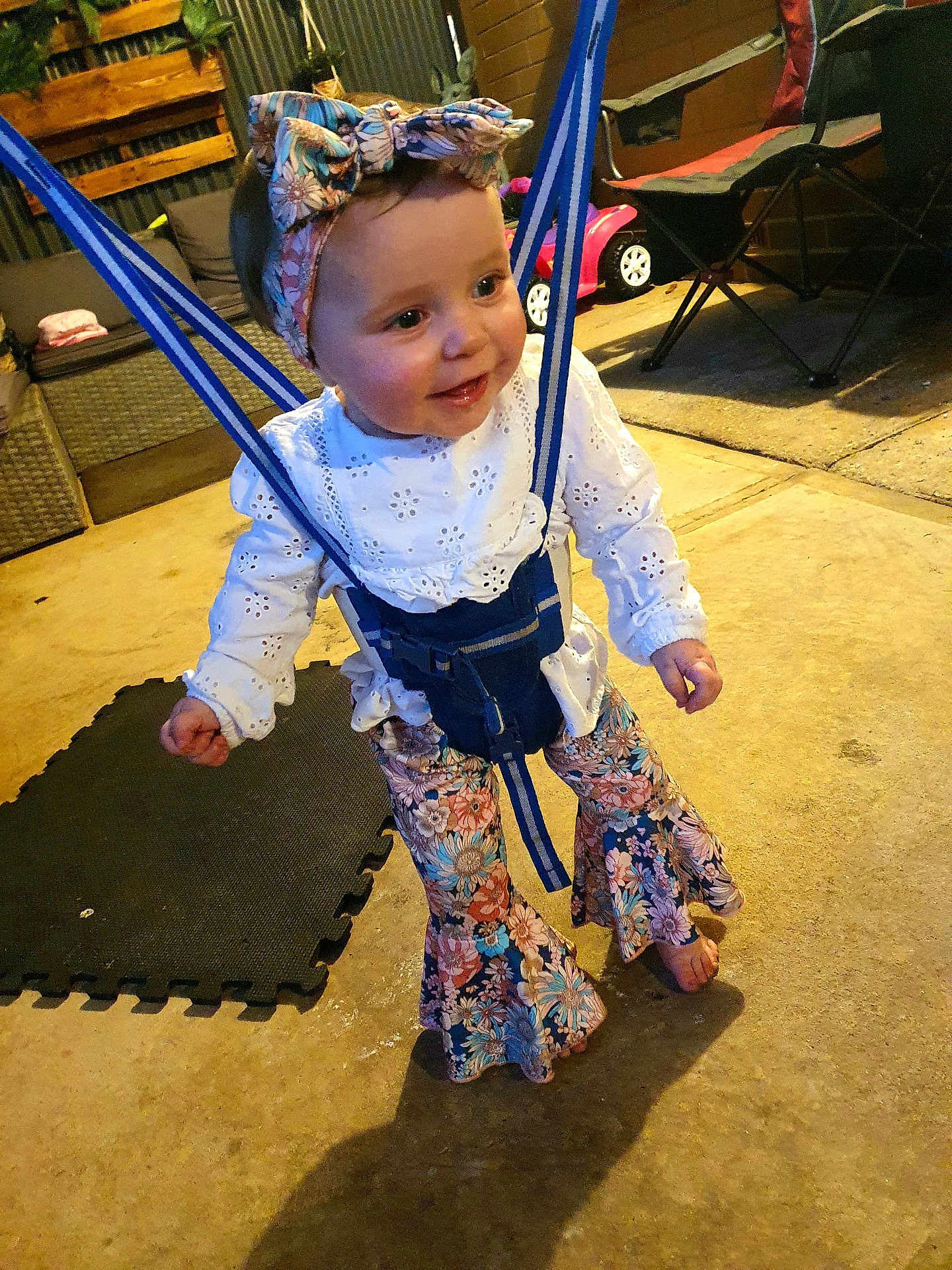 Reign joined the competition — help win amazing prizes! child, toddler, baby, harness, floral_headband, floral_pants, white_blouse, smiling, barefoot, play_area, toy_car, folding_chair, patio, wicker_sofa, foam_mat, concrete_floor, evening_light, portrait, happy, clothing