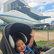 Andrew joined the competition — help win amazing prizes! airplane, baby, blue_shirt, child, clouds, daytime, fence, happy, infant, outdoor, park, pavement, seatbelt, sky, smiling, space_shuttle, stroller, technology, tourist_attraction, transportation