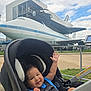 airplane, baby, blue_shirt, child, clouds, daytime, fence, happy, infant, outdoor, park, pavement, seatbelt, sky, smiling, space_shuttle, stroller, technology, tourist_attraction, transportation