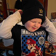 Joshua is registered to the contest to win money with this photo: beanie, boy, casual, child, clothing, cozy, expression, face, glasses, hat, indoor, mario_kart, person, playful, room, shirt, smile, table, toys, young