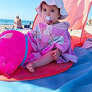 Inaya participe au concours pour gagner de l'argent avec cette photo : baby, beach, beach_mat, blue_sky, bucket, child, outdoor, pacifier, person, pink_towel, relaxation, sand, sea, summer, sunhat, sunny, sunshade, toddler, vacation, water