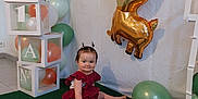 Inaya participe au concours pour gagner de l'argent avec cette photo : balloons, birthday, blocks, celebration, child, cute, decorations, deer_balloon, event, festive, floor, golden_balloon, green_carpet, indoors, party, red_dress, sitting, smiling, toddler, white_wall