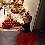 toddler, child, christmas_tree, holiday_decorations, red_tutu, santa_hat, tinsel, ornaments, lights, indoor, festive, holiday, white_tree, floor_tiles, brick_wall, cute, smiling, person, celebration, decor