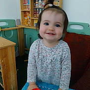 Inaya a rejoint le concours — aidez-le/la à gagner de superbes lots ! background, blue_eyes, child, cute, furniture, hair_tie, hands_on_table, happy, indoor, long_sleeve_shirt, patterned_clothing, person, playroom, red_box, smile, standing, table, toddler, toy_shelf, wooden_furniture