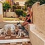 woman, dog, husky, white_dog, outdoor, courtyard, bench, plants, flowers, stone_wall, tile_floor, summer, sunlight, leash, pet, casual_clothing, smiling, portrait, nature, relaxing
