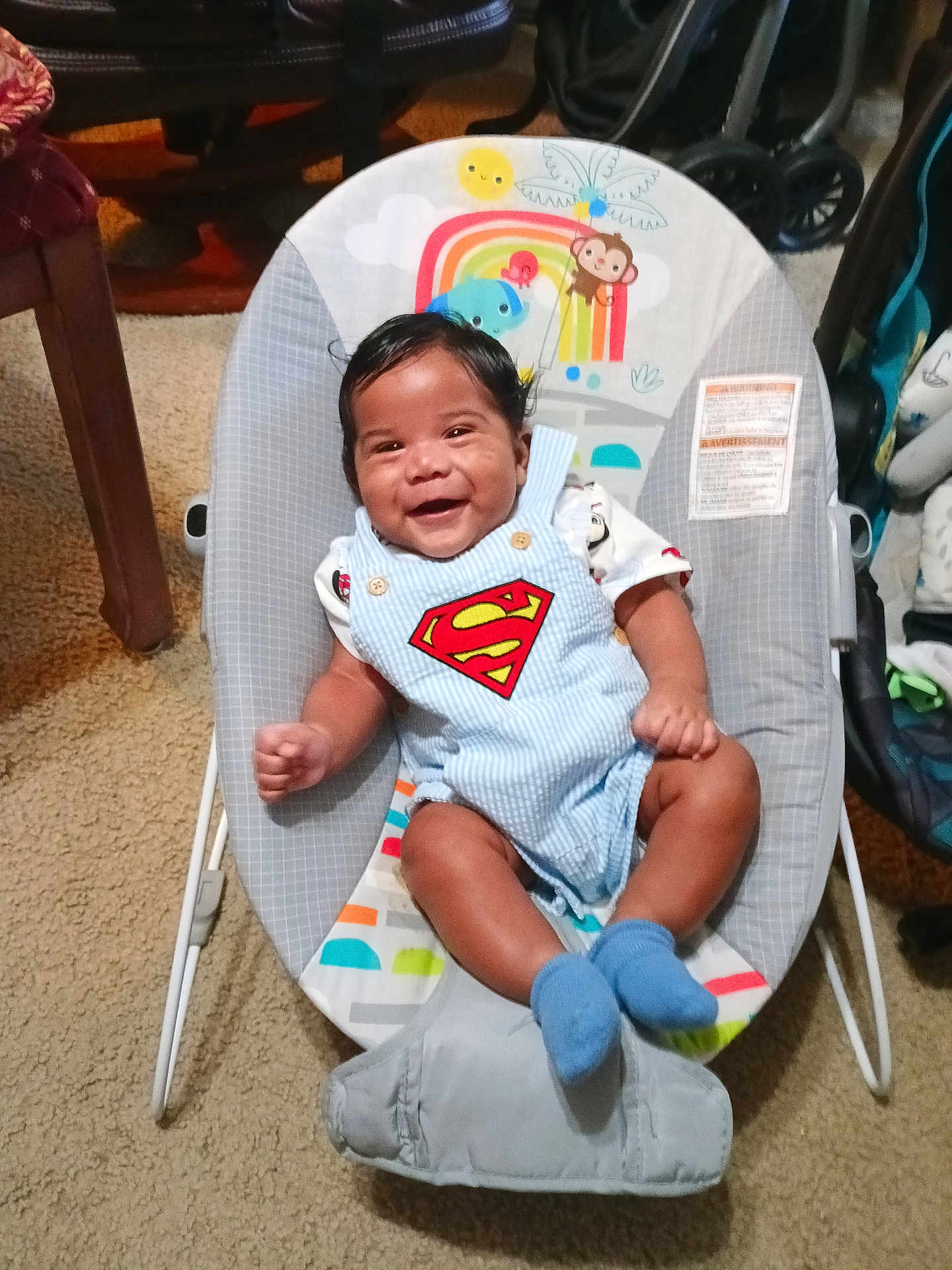 Kashton is registered to the contest to win money with this photo: baby, infant, smiling, romper, superman_logo, baby_bouncer, baby_seat, socks, rainbow, monkey, carpet, stroller, indoor, portrait, cute, happy, clothing, hair, furniture, living_room