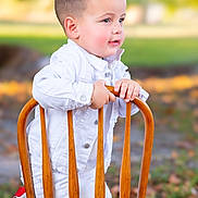 Dylan Michel is registered to the contest to win money with this photo: autumn_leaves, bracelet, casual, child, colorful_shoes, curious_expression, cute, daylight, fashion, green_background, kneeling, outdoor, playful, portrait, short_hair, smiling, toddler, white_clothing, wooden_chair, young_child