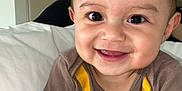 Jose joined the competition — help win amazing prizes! baby, smiling, infant, face, eyes, onesie, brown_clothing, yellow_print, bed, pillow, skin, hair, happy, cute, indoor, person, portrait, sitting, child, expression