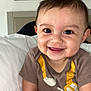 baby, smiling, infant, face, eyes, onesie, brown_clothing, yellow_print, bed, pillow, skin, hair, happy, cute, indoor, person, portrait, sitting, child, expression