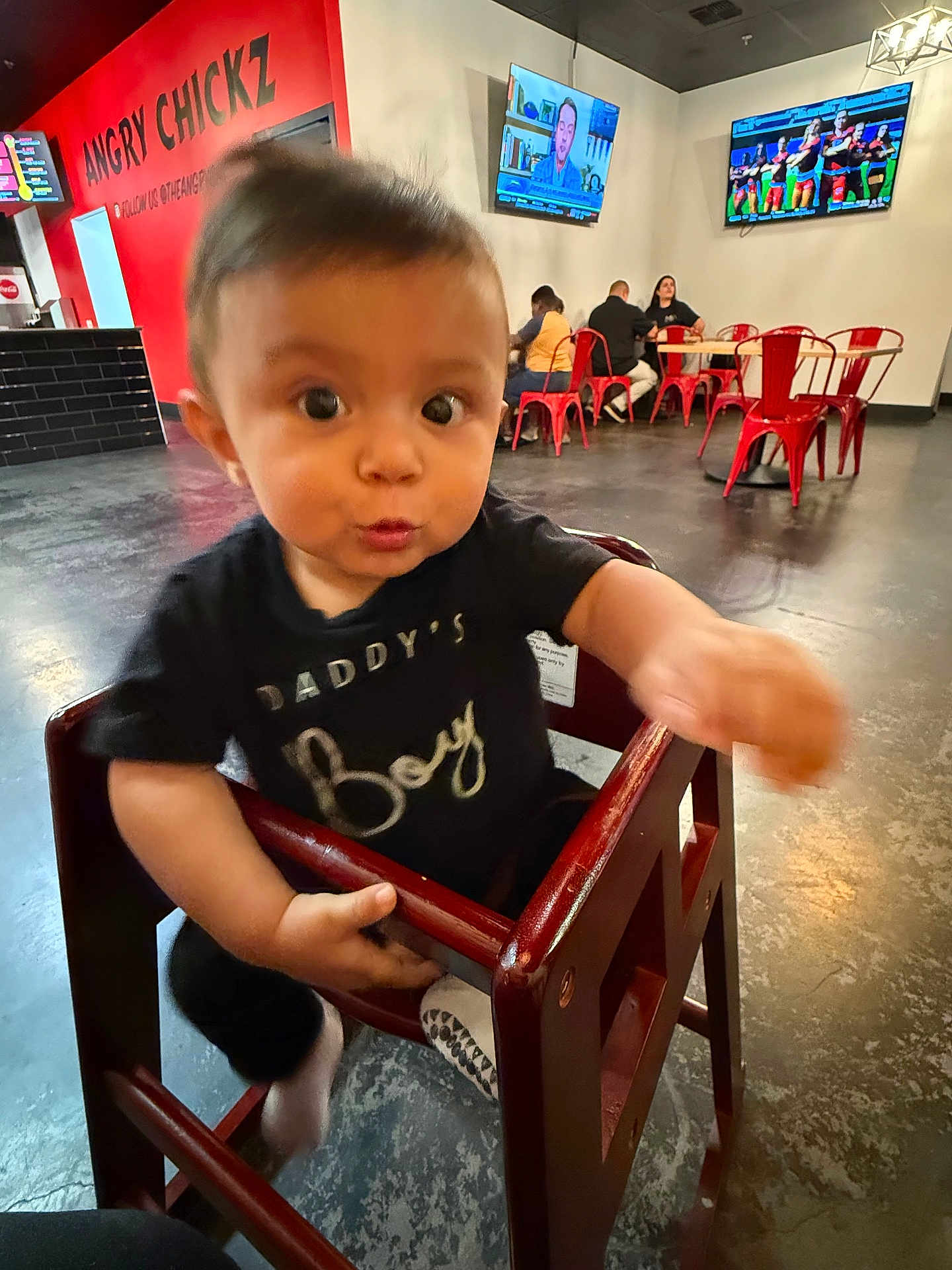 Jose is registered to the contest to win money with this photo: toddler, child, high_chair, restaurant, indoor, person, furniture, floor, television, table, chair, red_chair, black_tshirt, expression, face, hand, curious, playful, baby, people