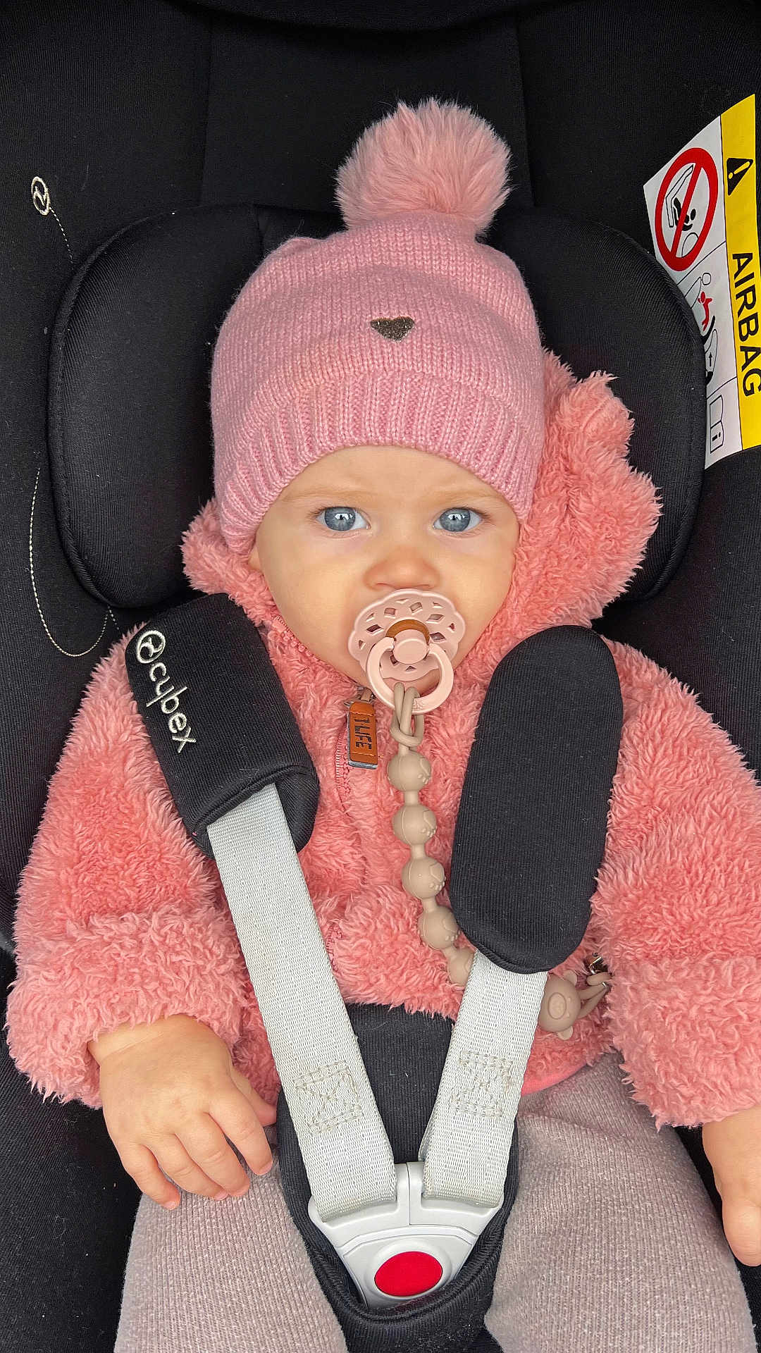 Lou participe au concours pour gagner de l'argent avec cette photo : baby, child, car_seat, pink_hat, pom_pom, pacifier, blue_eyes, fluffy_jacket, safety_harness, cute, infant, winter_clothing, warm, seatbelt, portrait, young_child, indoors, close_up, person, child_safety