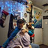 Fat Boy is registered to the contest to win money with this photo: person, dog, hugging, indoor, blue_hoodie, purple_shirt, baby_yoda, plush_toy, stocking, poster, blue_lights, chair, door, wall, floor, glasses, hand, furniture, affection, casual