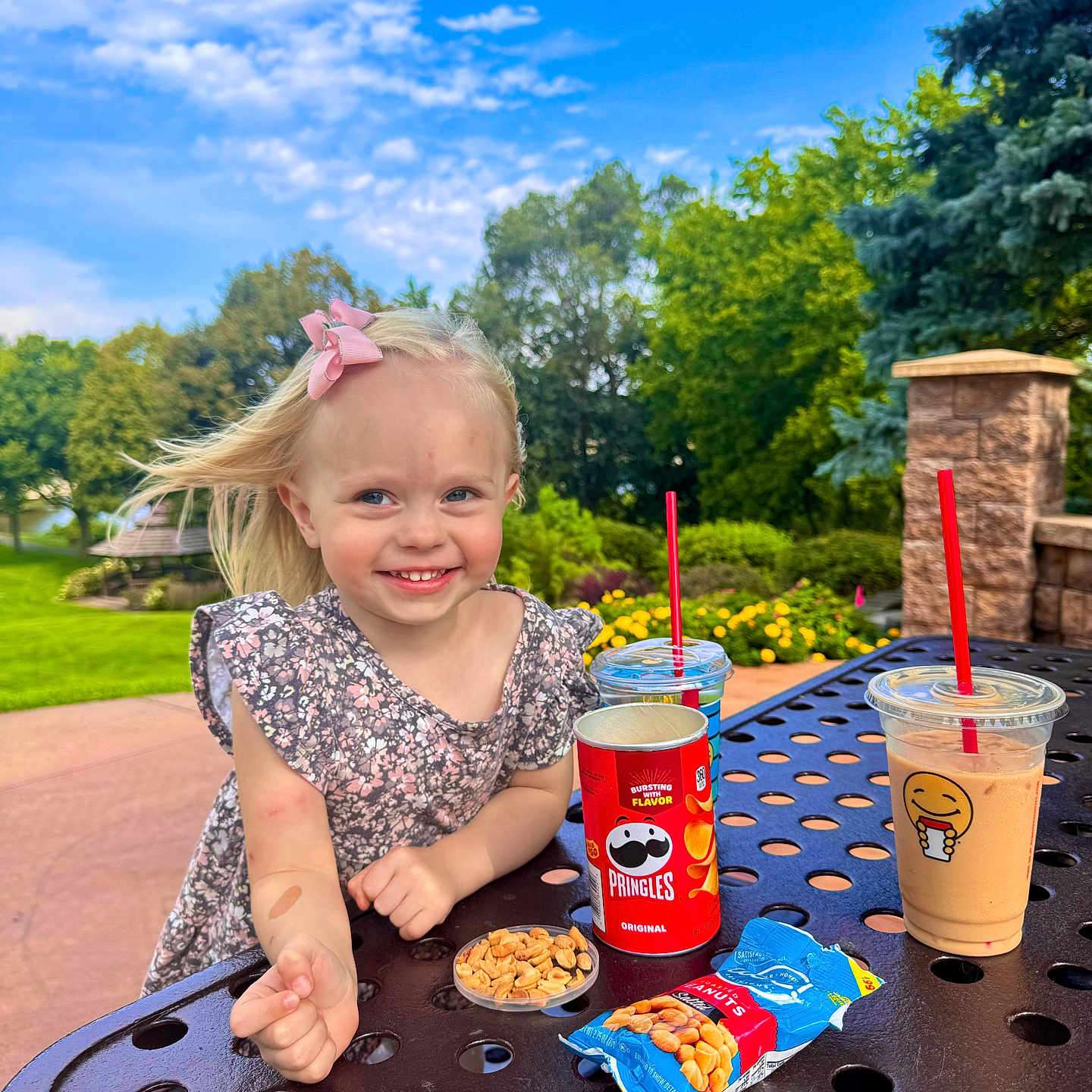 Amelia is registered to the contest to win money with this photo: blue_sky, child, clouds, daytime, dress, girl, greenery, happy, iced_drinks, outdoor, park, peanuts, picnic_table, pink_bow, pringles, red_straw, smiling, snacks, summer, trees