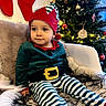 celebration, chair, child, christmas_elf, christmas_tree, cozy, cute, decorations, festive, furry_blanket, happy, hat, holiday, holiday_outfit, indoor, smiling, stockings, striped_pants, toddler, young_child