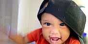 Amirr is registered to the contest to win money with this photo: baby, blurred_background, cap, casual_clothing, child, cute, expression, face, hand, happy, headwear, holding, indoor, infant, motion_blur, orange_shirt, person, playful, portrait, smiling