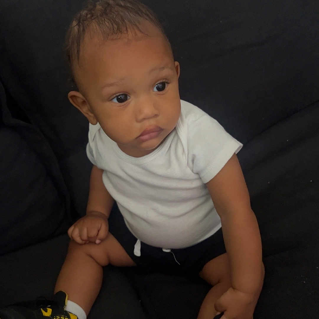 Amirr is registered to the contest to win money with this photo: baby, black_background, black_shorts, black_yellow_shoes, casual_clothing, child, couch, curious_expression, footwear, indoor, infant, looking_away, person, seated, sitting, small_hands, sneakers, toddler, white_tshirt, young_child