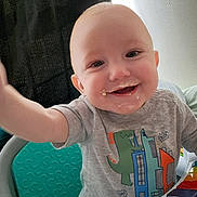 Ryland is registered to the contest to win money with this photo: baby, child, smiling, high_chair, food_mess, dinosaur_shirt, happy, face, reaching_hand, indoor, toy, colorful, person, toddler, playful, cute, clothing, seat, table, infant