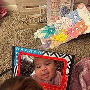 Eden is registered to the contest to win money with this photo: baby, reflection, mirror, carpet, hair_bows, wooden_crate, toy, colorful, child, indoor, face, curly_hair, fabric, pink, orange, blue, purple, decor, playtime, closeup