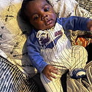 Messiah is registered to the contest to win money with this photo: baby, infant, child, portrait, indoors, lying_down, blanket, pillows, couch, sleepwear, onesie, blue_clothing, baseball_theme, striped_clothing, dark_skin, black_hair, face, eyes, casual, home