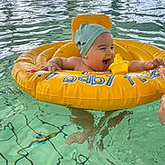 Selyan participe au concours pour gagner de l'argent avec cette photo : baby, infant, swimming_pool, inflatable_ring, water, child, indoor_pool, swim_cap, floating, happy, reaching, people, background, windows, natural_light, yellow, blue, splash, playful, fun