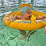 baby, infant, swimming_pool, inflatable_ring, water, child, indoor_pool, swim_cap, floating, happy, reaching, people, background, windows, natural_light, yellow, blue, splash, playful, fun