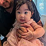 baby, child, adult, person, pigtails, pink_dress, golden_accents, holding, hand, face, eyes, smile, indoor, window_blinds, natural_light, curly_hair, cute, soft_fabric, toddler, family