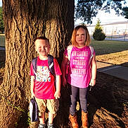 Olivia is registered to the contest to win money with this photo: backpack, boots, boy, building, casual_clothing, child, daytime, expression, girl, grass, nature, outdoor, park, pink_shirt, shadow, shorts, sidewalk, standing, sunlight, tree