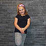 balance, black_shirt, brick_wall, casual_clothing, child, fashion, full_body, girl, gray_leggings, happy, headband, indoor, pink_bow, playful, portrait, posing, smiling, standing, young, youth