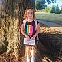 Olivia is registered to the contest to win money with this photo: backpack, casual_clothing, child, daylight, girl, glasses, grass, happy, nature, outdoor, paper, park, portrait, shoes, sidewalk, smile, summer, sunlight, tree, young_person