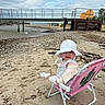 baby, beach, sand, chair, hat, child, water, pier, rocks, cloudy_sky, person, shore, outdoor, coast, footwear, clothing, pink, relaxation, summer, nature