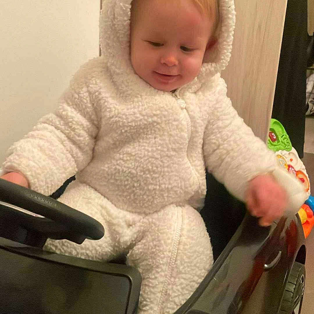 Leyten a rejoint le concours — aidez-le/la à gagner de superbes lots ! baby, car_toy, child, cute, face, floor, furniture, hand, head, indoor, onesie, play, plush, seat, smiling, steering_wheel, toddler, toy_car, white_clothing, wood_panel