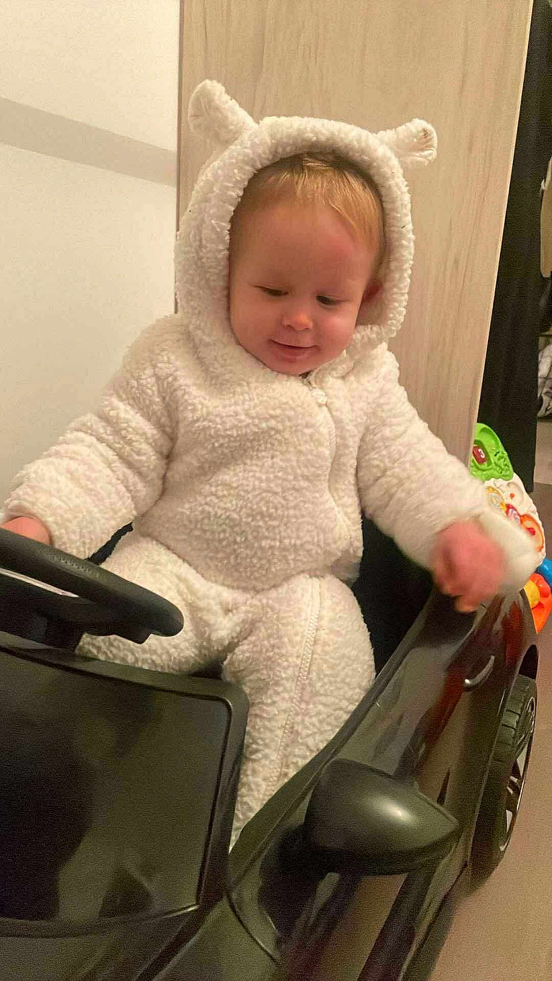 Leyten a rejoint le concours — aidez-le/la à gagner de superbes lots ! toddler, child, toy_car, onesie, white_clothing, indoor, cute, smiling, baby, plush, seat, steering_wheel, car_toy, floor, face, head, hand, furniture, wood_panel, play
