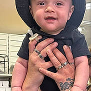 Draco joined the competition — help win amazing prizes! baby, child, cowboy_hat, portrait, smiling, happy, infant, held, hands, pacifier, black_clothing, tattoo, rings, indoor, kitchen, background, close_up, face, text_overlay, photography