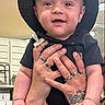 baby, child, cowboy_hat, portrait, smiling, happy, infant, held, hands, pacifier, black_clothing, tattoo, rings, indoor, kitchen, background, close_up, face, text_overlay, photography