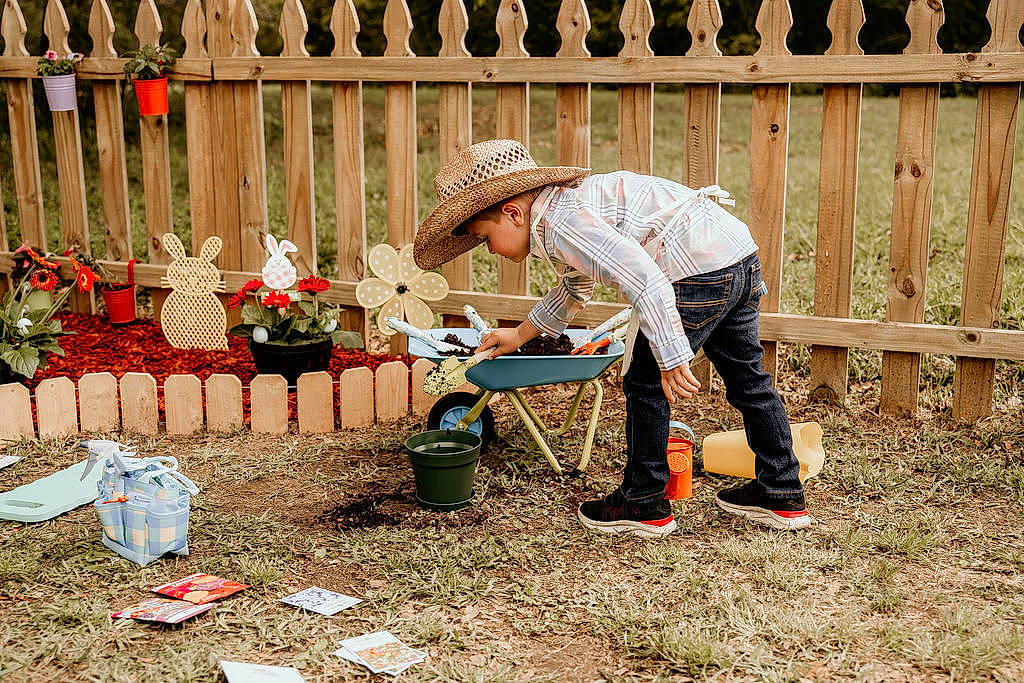 Preston is registered to the contest to win money with this photo: child, fence, flooring, fun, garden, gardening, grass, hat, home_fencing, landscape, landscaping, lawn, leisure, people_in_nature, person, plant, recreation, sitting, soil, sun_hat