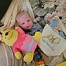 baby, infant, child, face, eyes, lying_down, smiling, blanket, bed, plush_toy, stuffed_animal, doll, duck_toy, pink, yellow, floral, bag, clothing, indoor, portrait