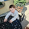 child, toddler, baby, outdoor, patio, chair, table, plant, flowerpot, window, house, courtyard, sunlight, shadow, portrait, sitting, leaning, floral_shirt, brown_hair, curious