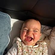 Gioia a rejoint le concours — aidez-le/la à gagner de superbes lots ! baby, infant, smile, laughing, face, teeth, bib, floral_bib, pillow, bed, headboard, plush_toy, blanket, joyful, cute, portrait, cheeks, eyes_closed, skin, indoor