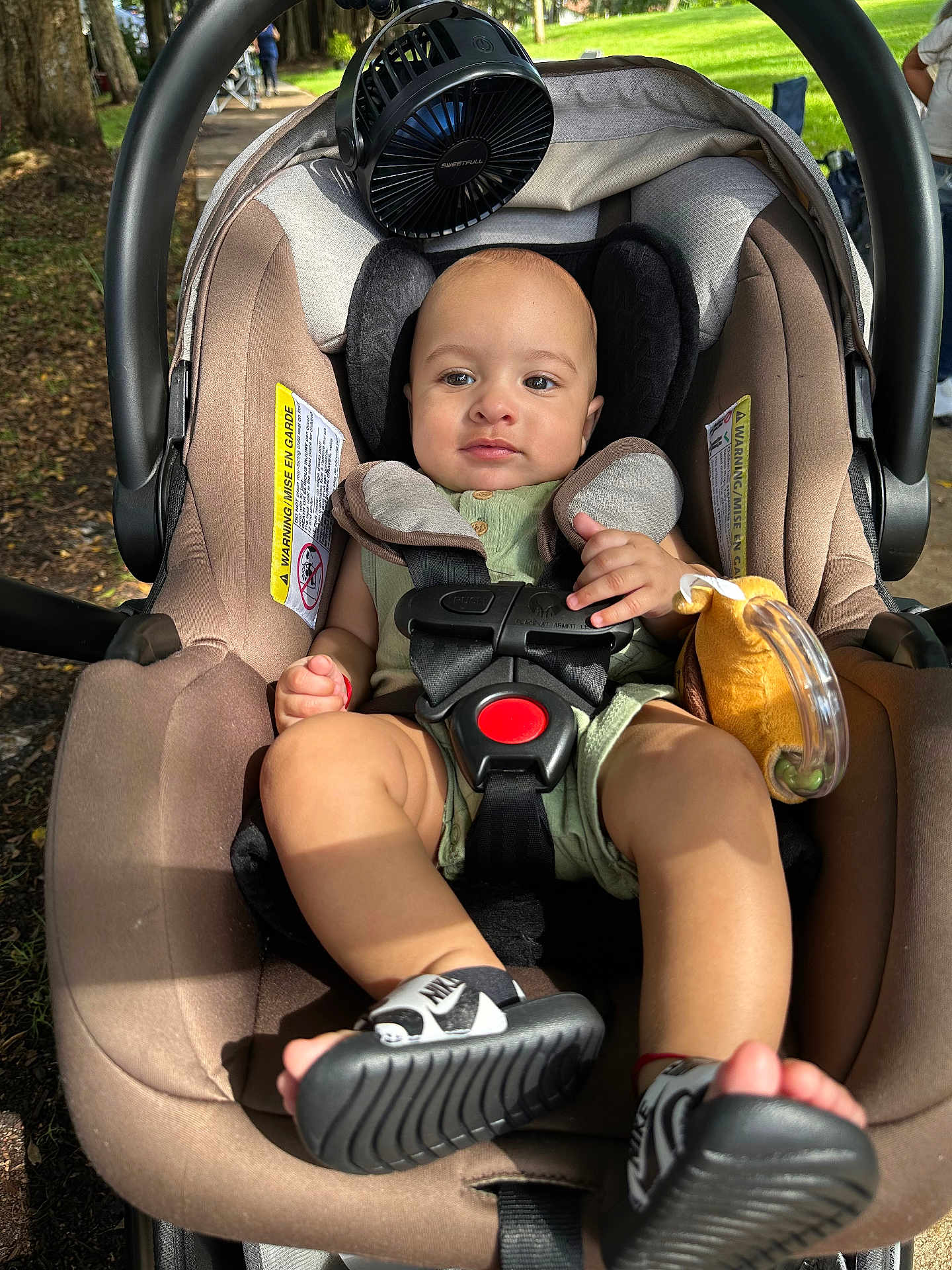 Liam is registered to the contest to win money with this photo: baby, car_seat, child, outdoor, greenery, toy, fan, footwear, nike, straps, sunlight, park, trees, person, infant, clothing, sitting, shoes, cute, summer
